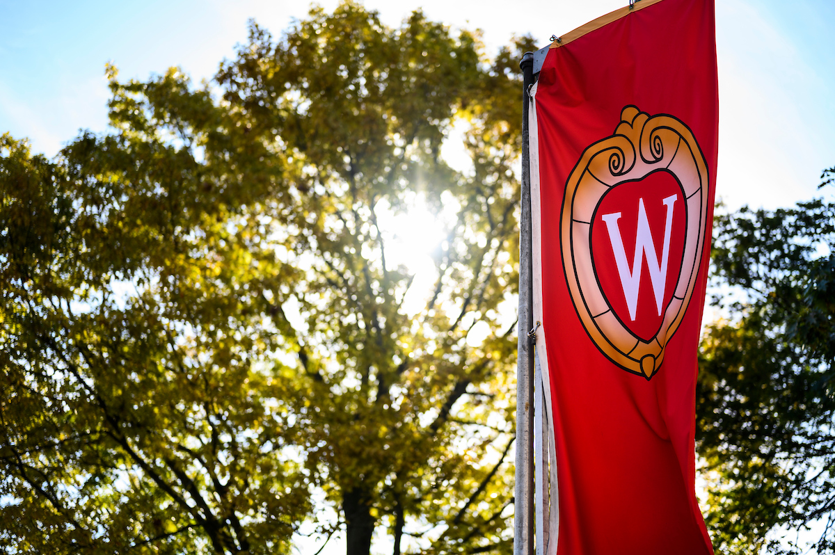 A red W crest banner flutters in the wind on Bascom Hill against a backdrop of trees and sunlight