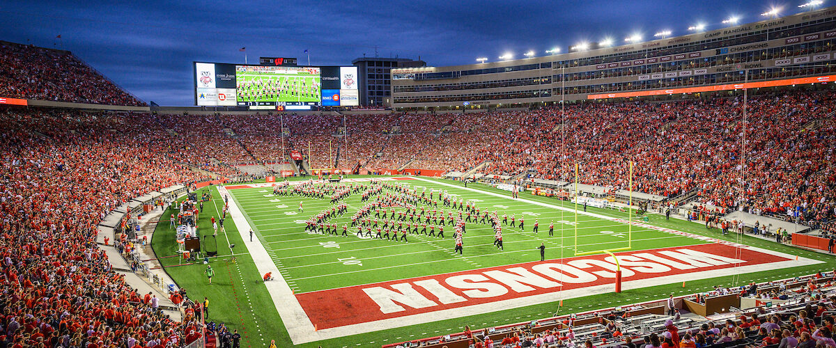 The UW Marching band performs in formation at a packed Camp Randall brightly illuminated by stadium lights under a darkening sky